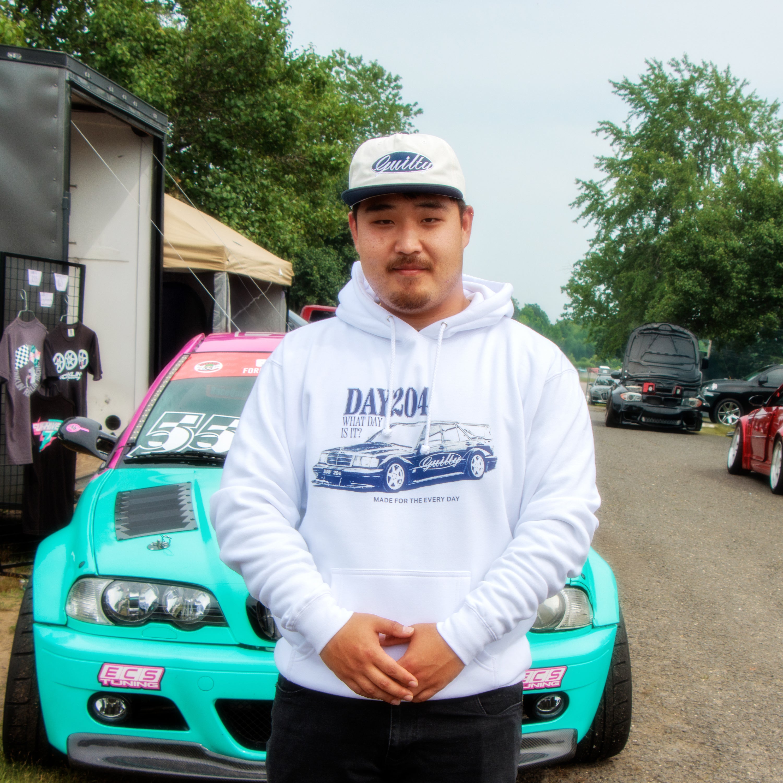 Man wearing a white hoodie with a car graphic and cap, standing in front of a turquoise car at an outdoor event.