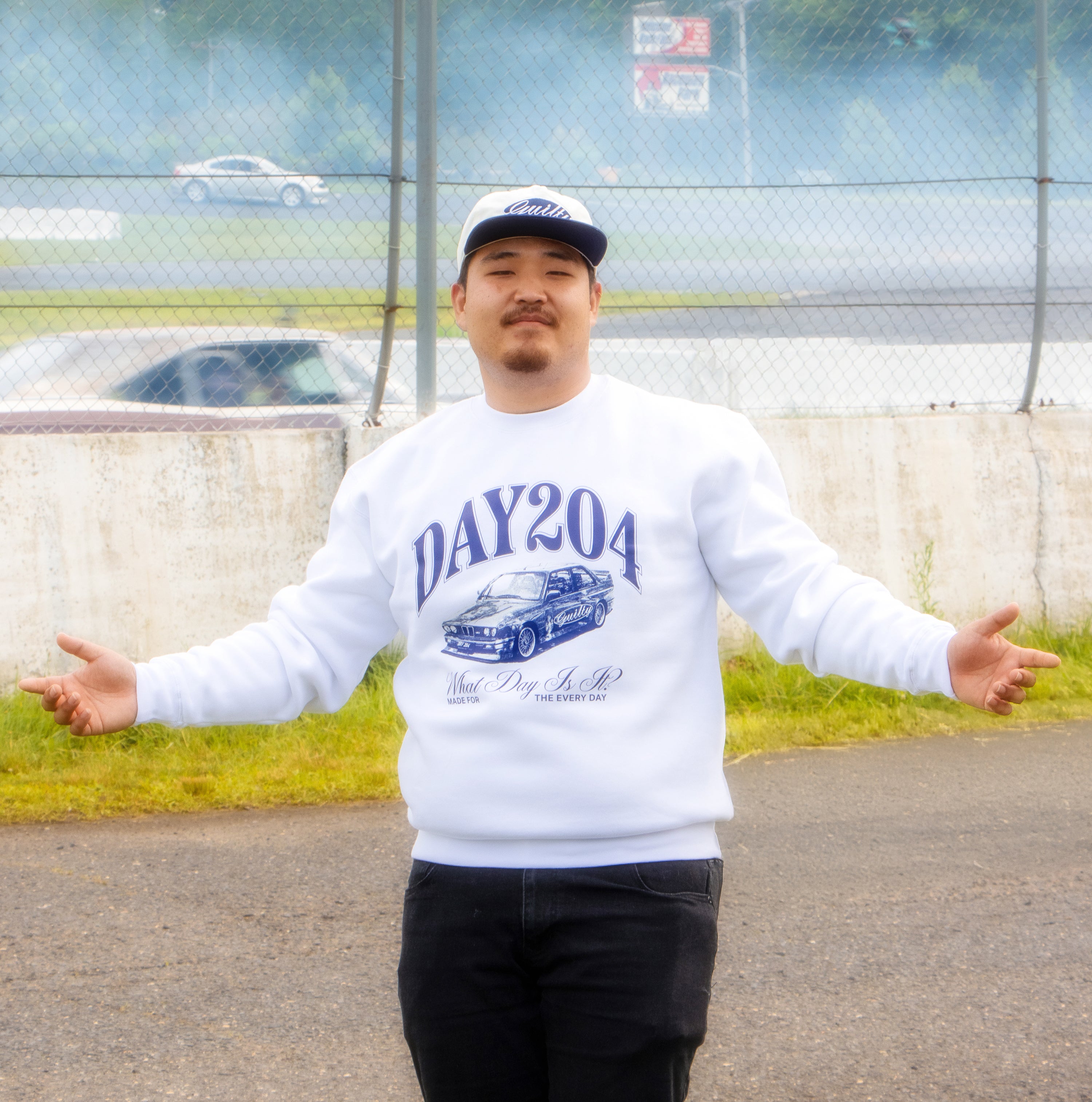 Person wearing a white sweatshirt with 'DAY204' print, standing outdoors near a fence.