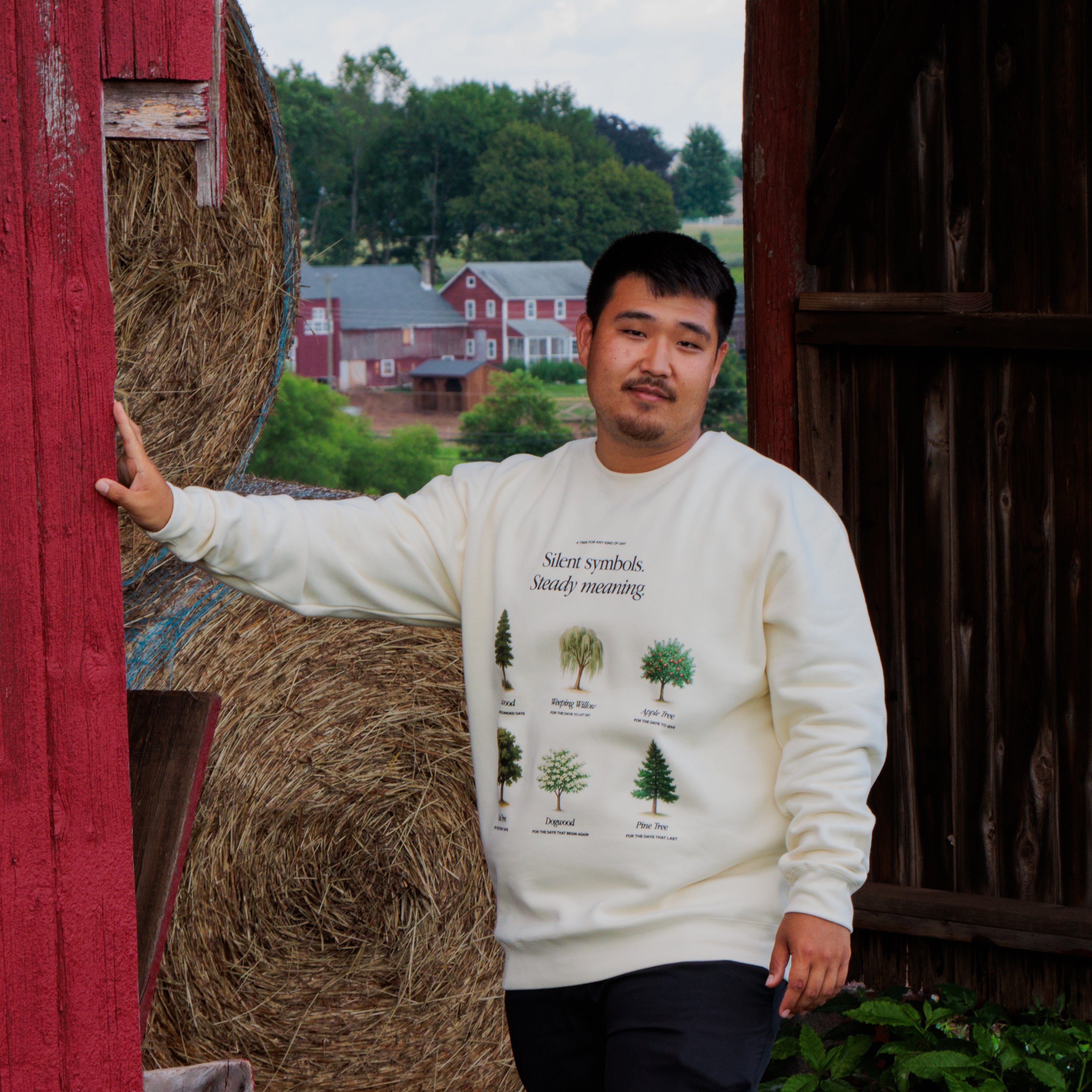 Man wearing a white sweatshirt with tree illustrations standing next to hay bales in a barn setting.