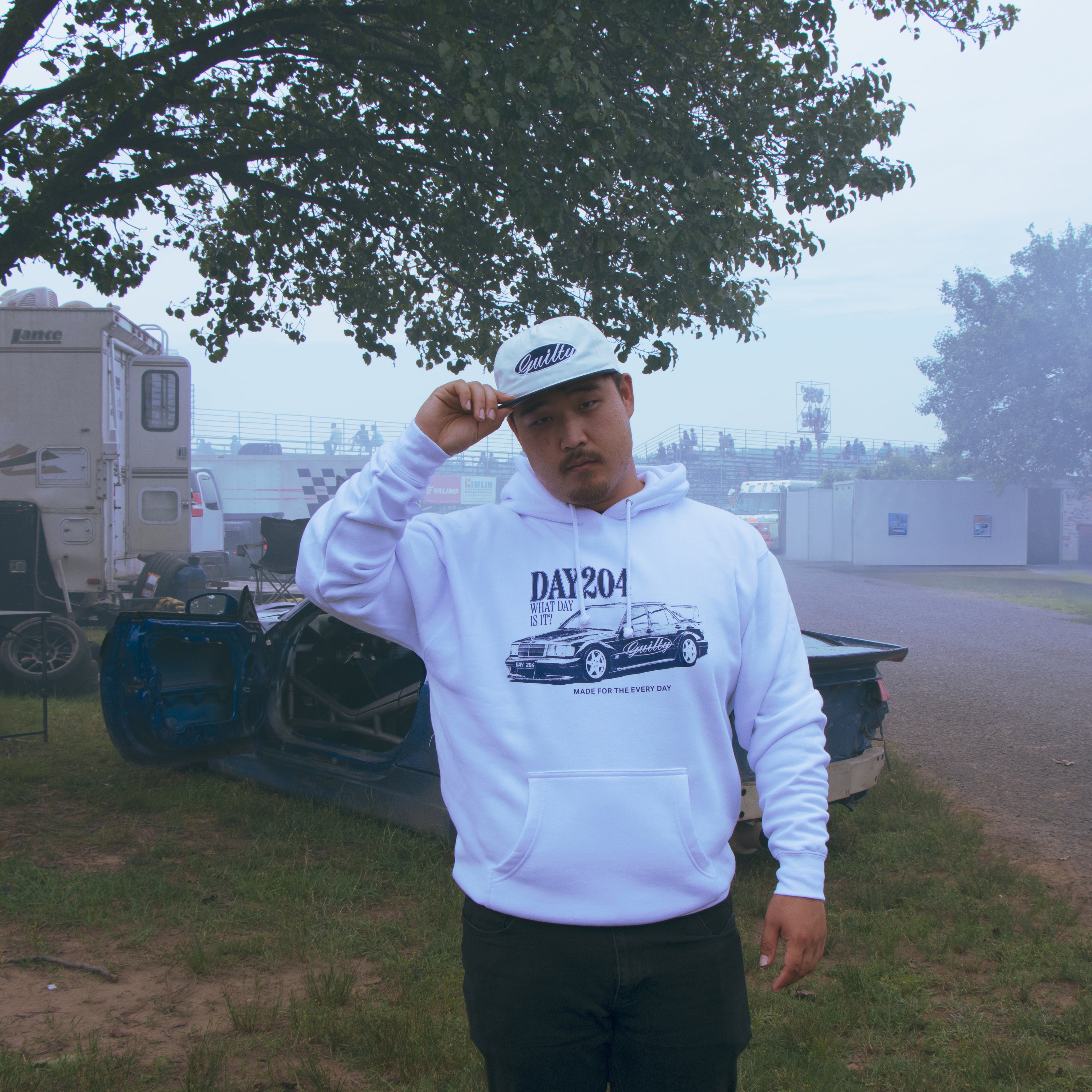 Person wearing a white hoodie with a graphic design, standing outdoors near trees and a trailer.