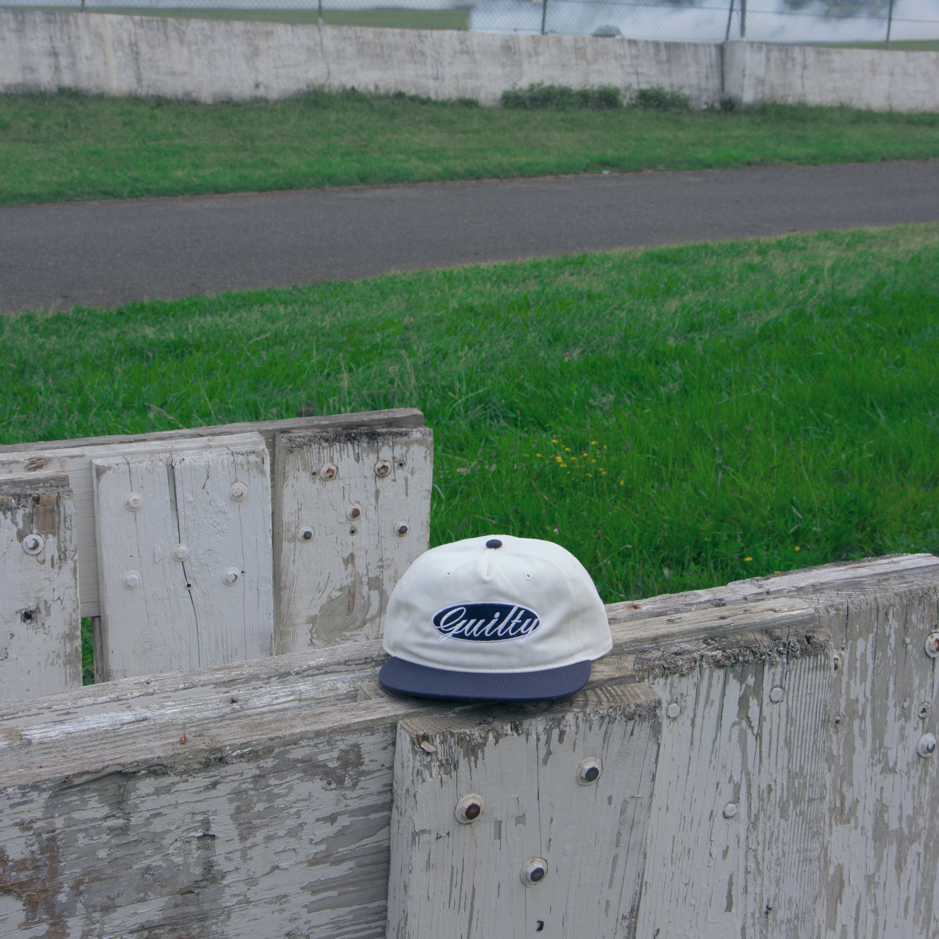 White cap with blue brim and 'Guilty' logo on a wooden fence with grass and road in the background