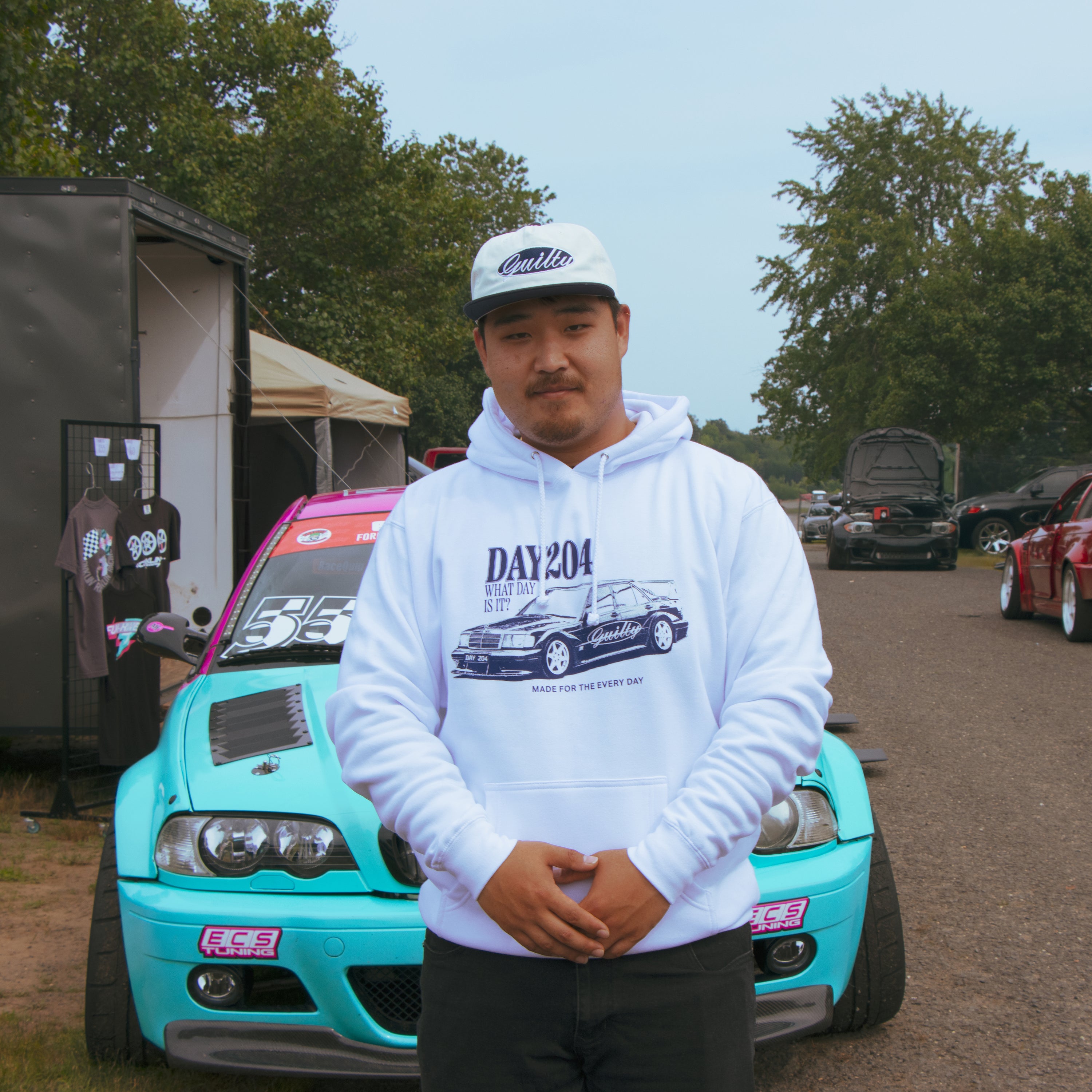 Man wearing a white hoodie with a car graphic in front of a turquoise car at an outdoor event.