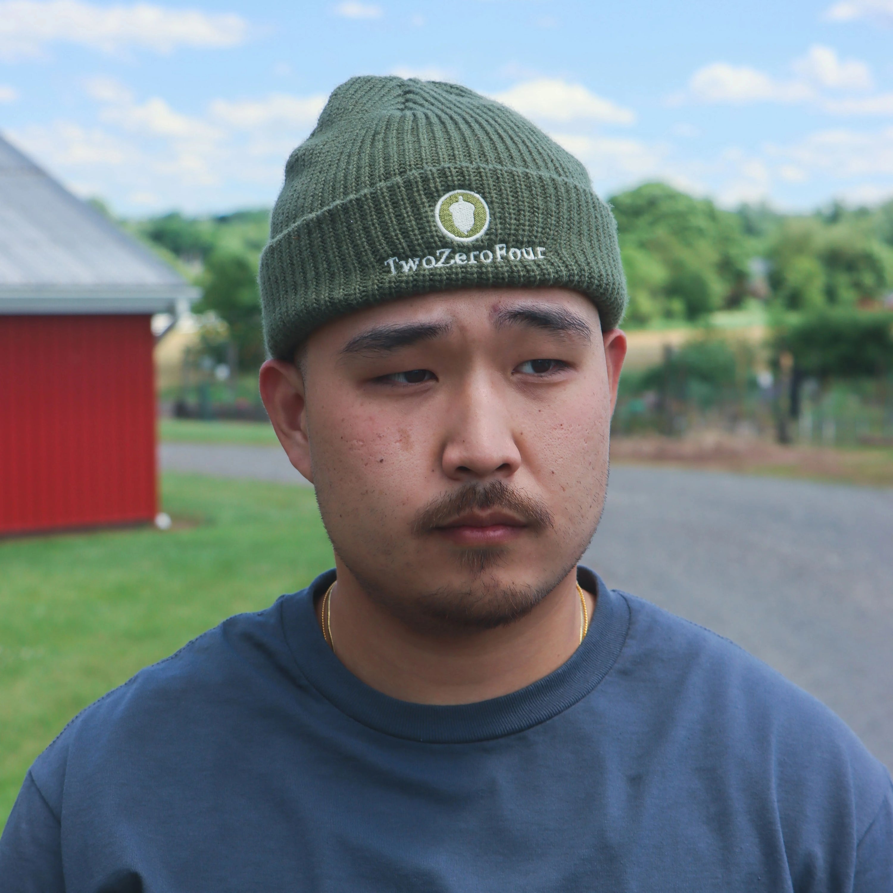 Man wearing a green beanie with a logo and text, standing outdoors with a red building and trees in the background.