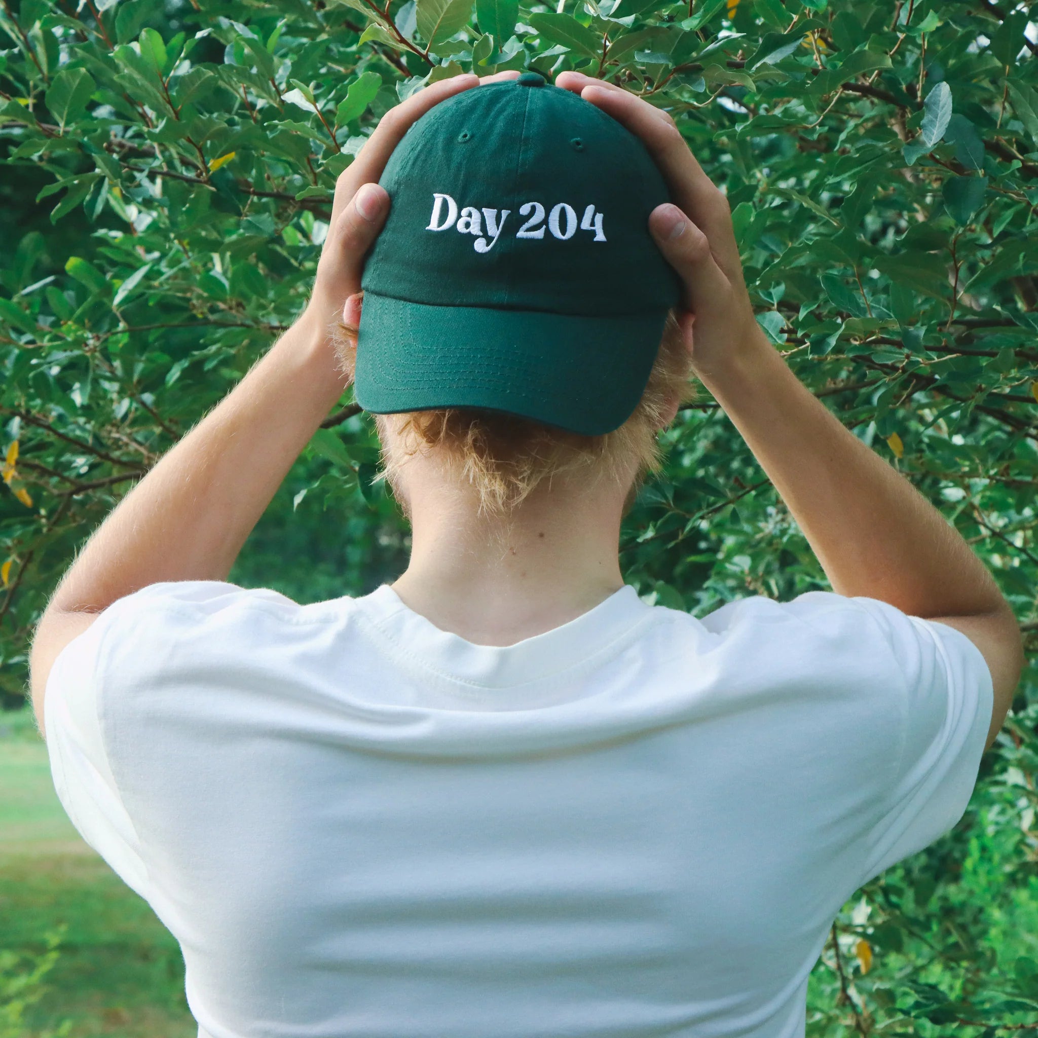 Person wearing a green cap with 'Day 204' text, standing in front of green foliage.