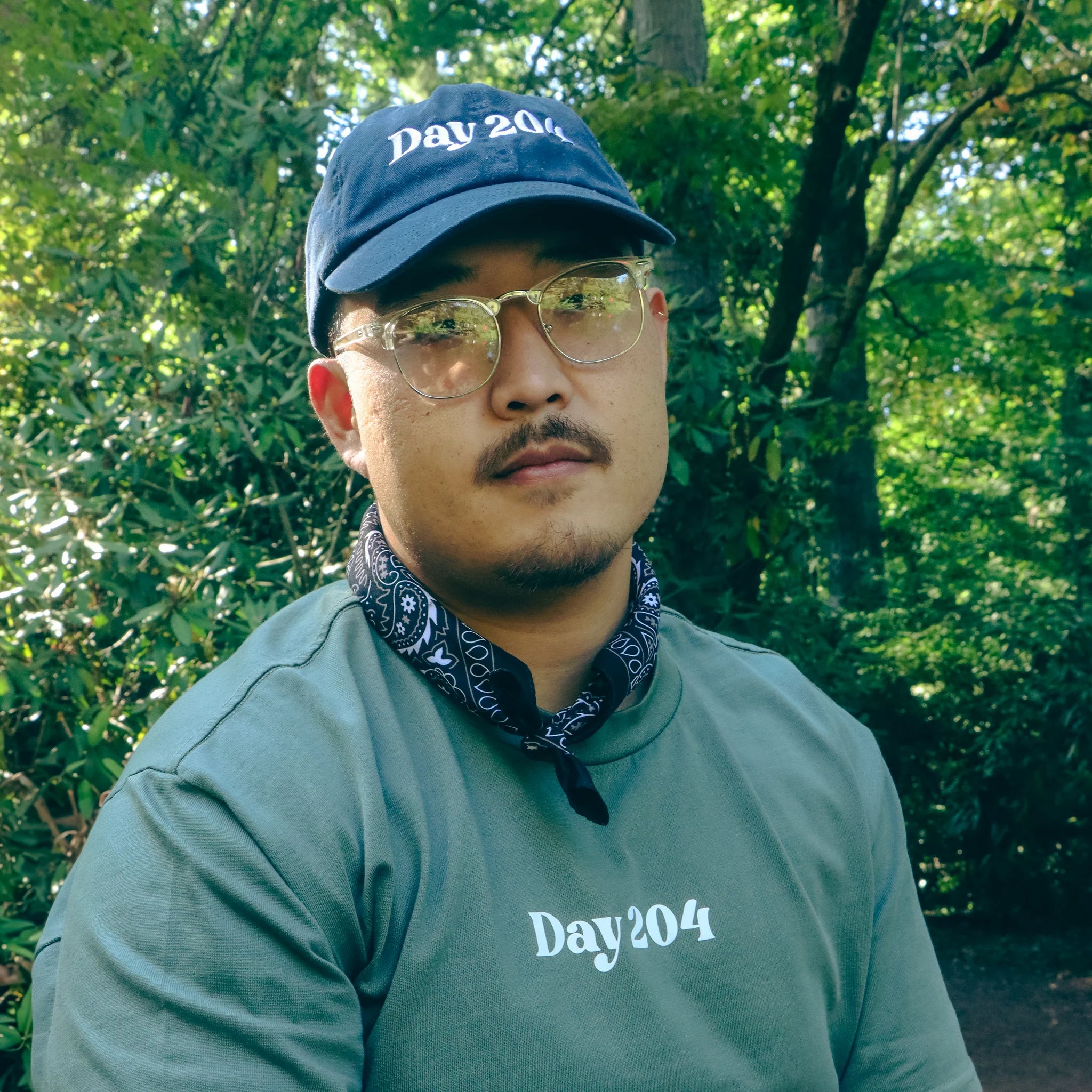 Man wearing a cap and sunglasses with a green shirt and bandana in a forest setting