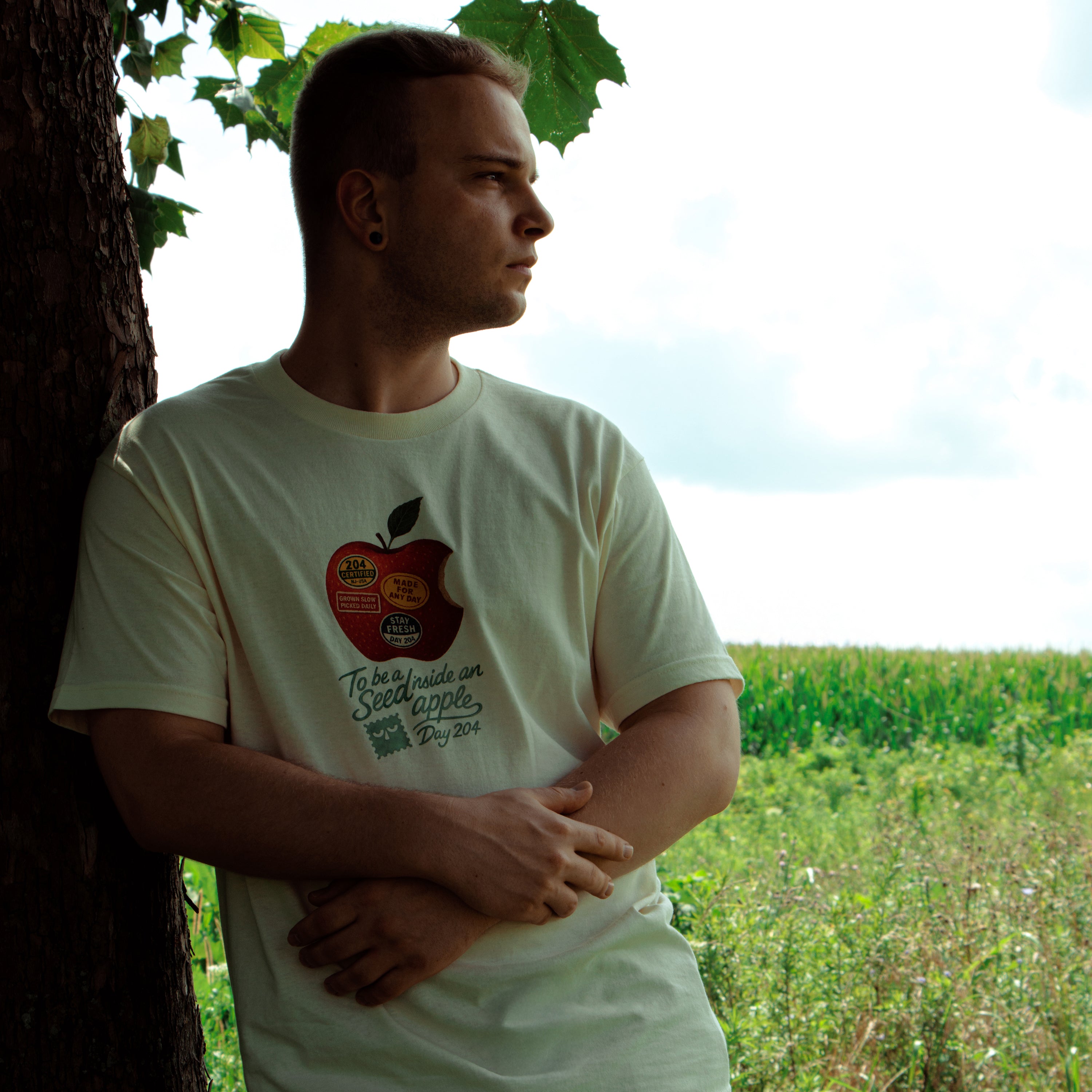 Man wearing a white t-shirt with an apple graphic and text, standing in a field.