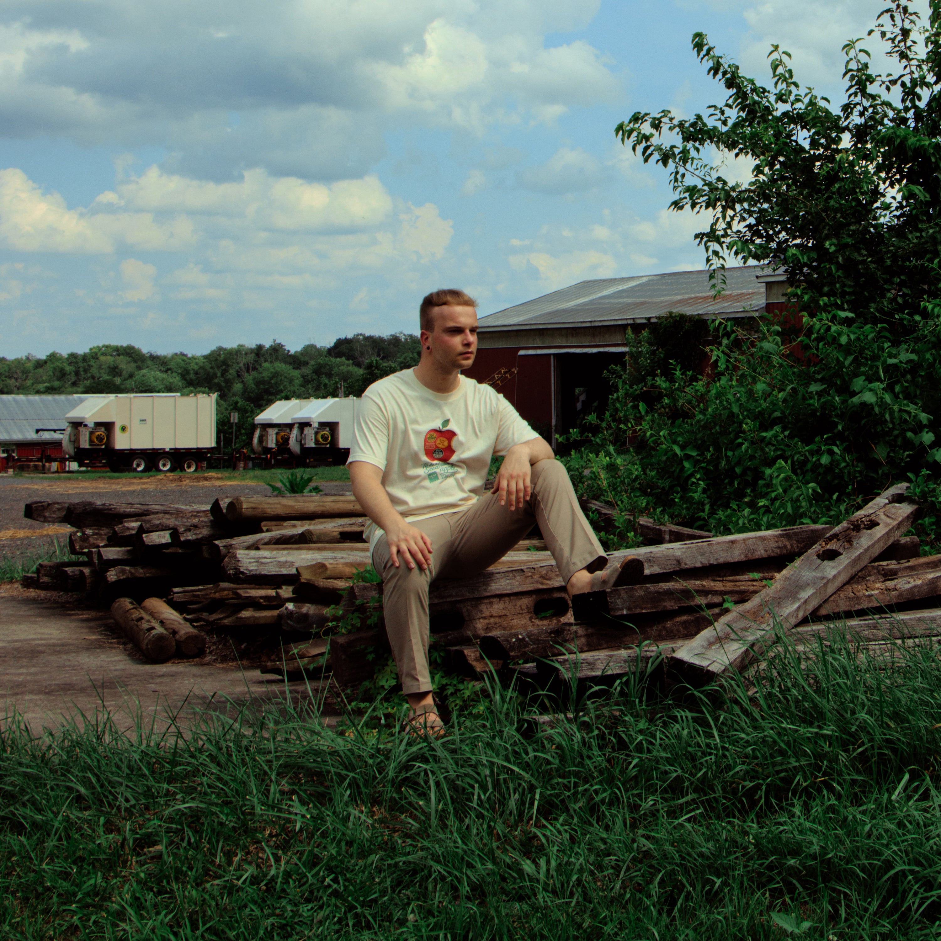 Man sitting on wooden planks in a rural setting with trees and a cloudy sky.